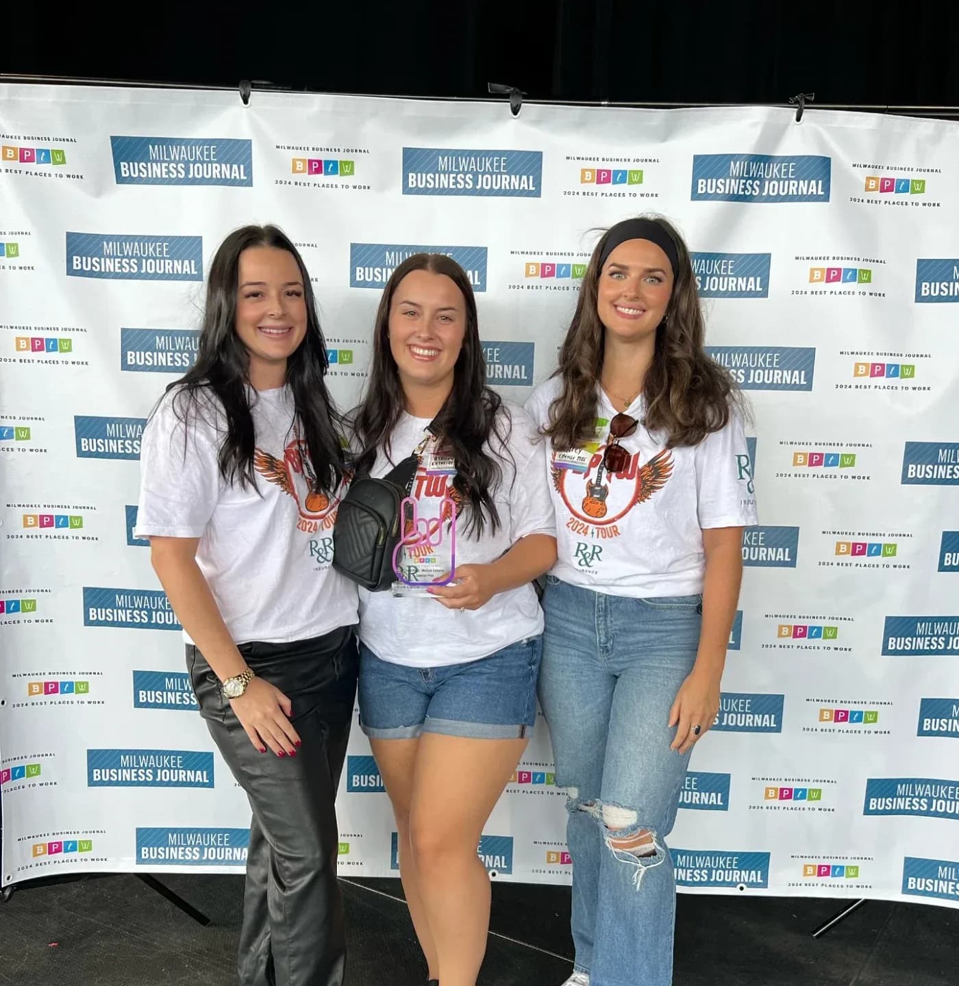 Three women posing at Milwaukee Business Journal Best Places to Work event backdrop, holding an award, wearing matching white graphic t-shirts