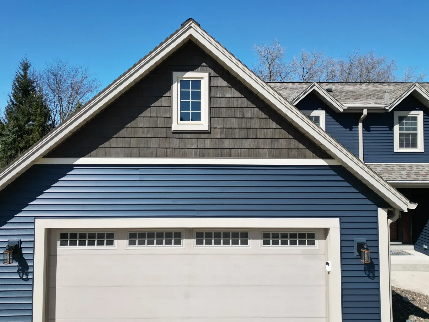 Exterior front view of a multi-story house featuring dark blue horizontal siding, dark grey shake siding in the upper gable peak, and white trim accents. A white double-car garage door with small top-row windows is framed by outdoor wall sconces, with a small rectangular window set in the upper gable under a clear blue sky.