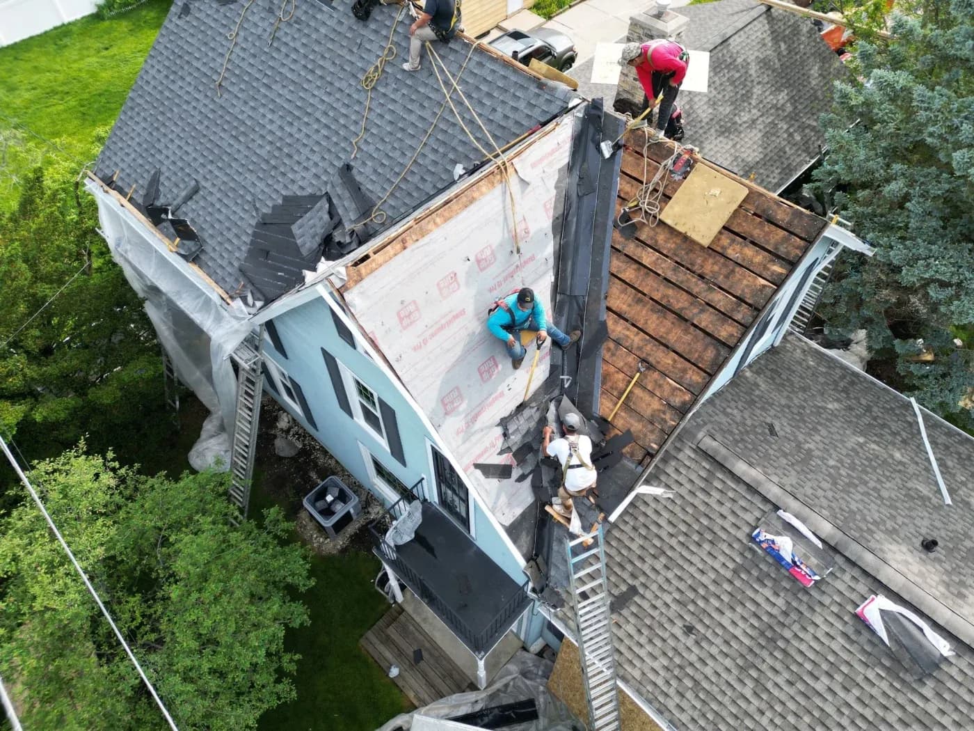 Aerial view of roofers installing shingles on a residential house, with workers secured by safety ropes working on the pitched roof and roof decking visible