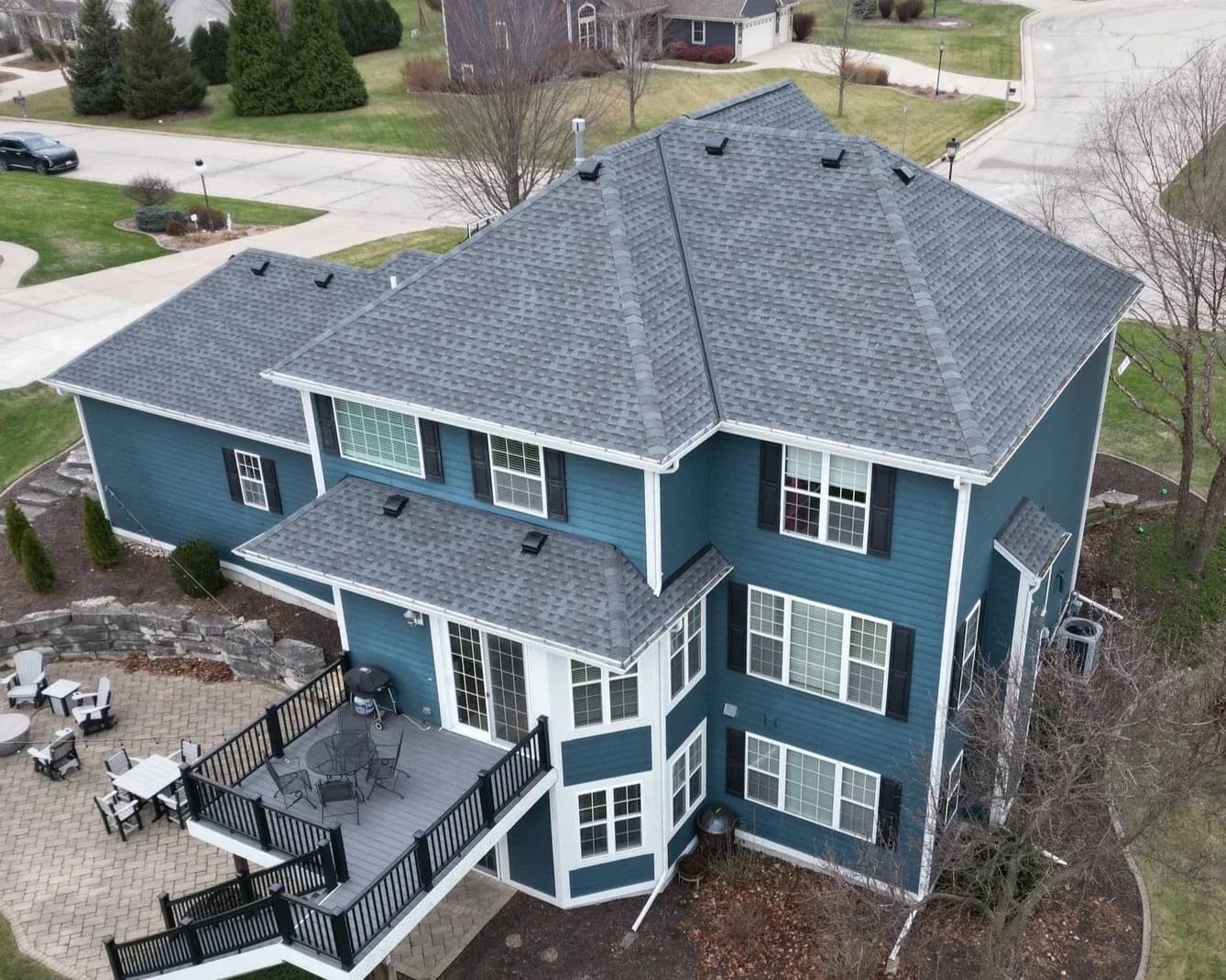 Aerial view of a two-story blue and white house with gray shingle roof, multiple windows, and a back deck with black railing overlooking a patio in a residential neighborhood