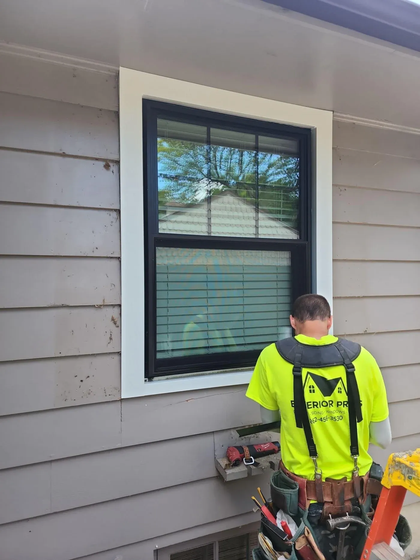 Worker in neon safety vest installing black double-hung window with white trim on gray siding house