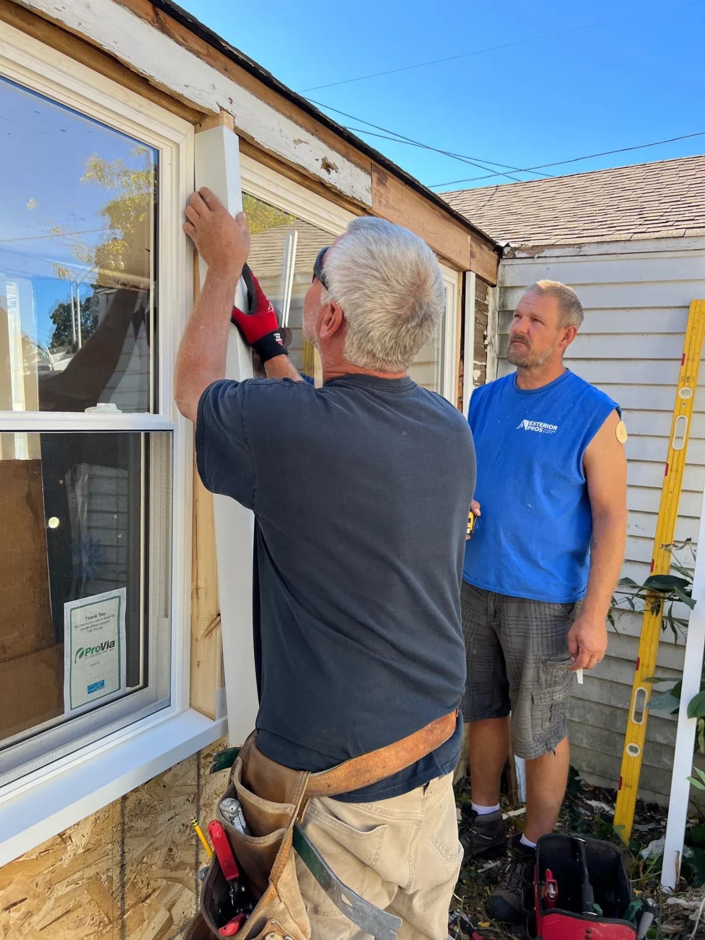 Two workers installing a window on a house exterior, with one man measuring the frame while another supervises the job