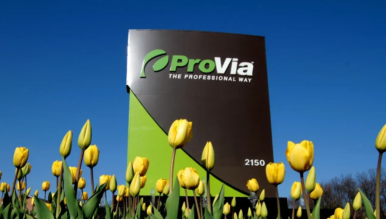 ProVia company sign with green and black design against blue sky, surrounded by yellow tulips in foreground