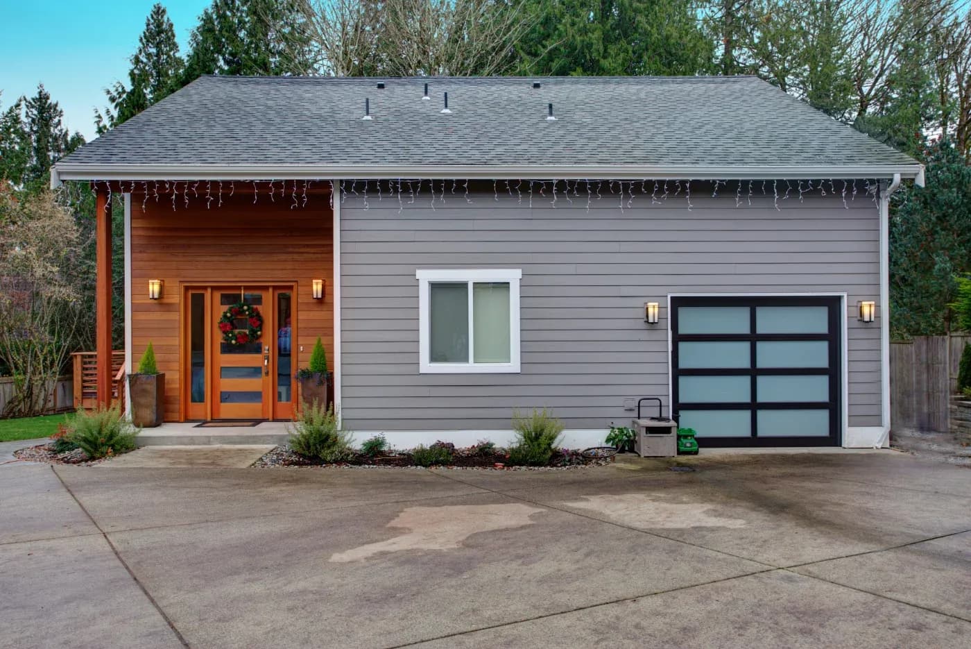 Exterior front view of a modern single-story home featuring grey horizontal lap siding and warm wood paneling around the entrance area. The entryway has a matching wooden front door decorated with a festive wreath and flanked by wall sconces. To the right, a white-framed sliding window sits on the main facade next to a modern black-framed garage door with frosted glass panels, set against a wide concrete driveway.