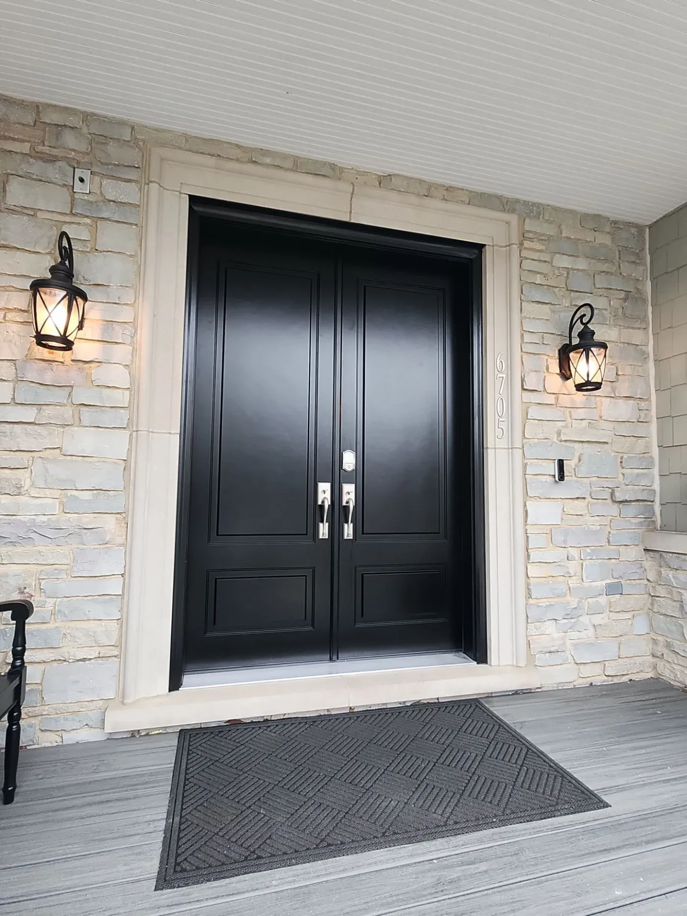 Black double entry doors with sidelights on stone brick exterior, flanked by lantern sconces and doormat on covered porch