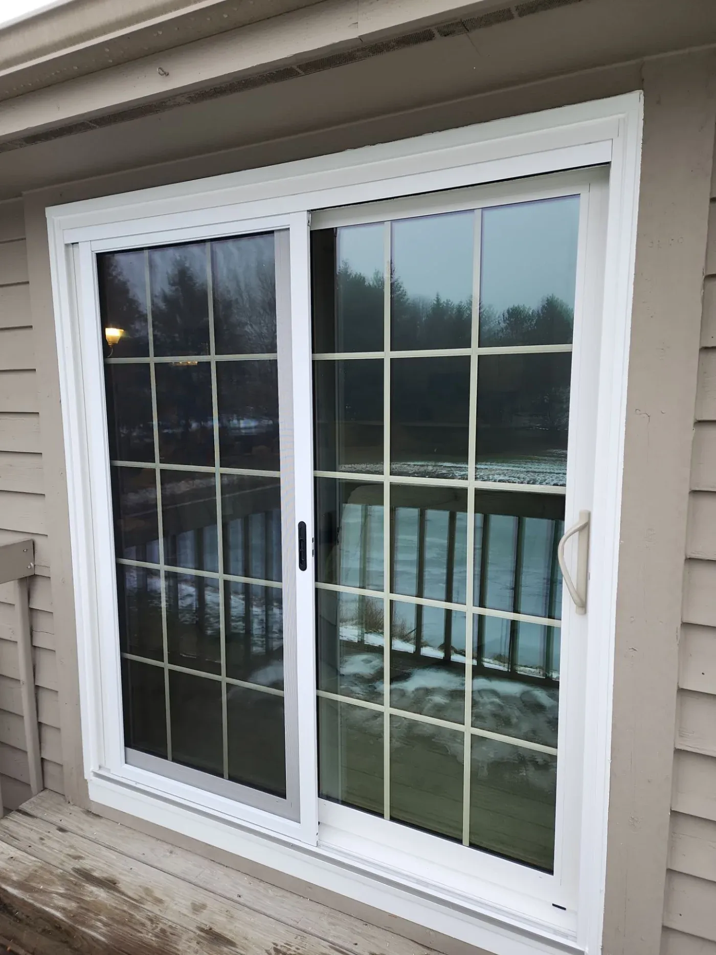 White-framed sliding glass patio doors with 15-pane grids opening onto a wooden deck overlooking a forested lake at dusk
