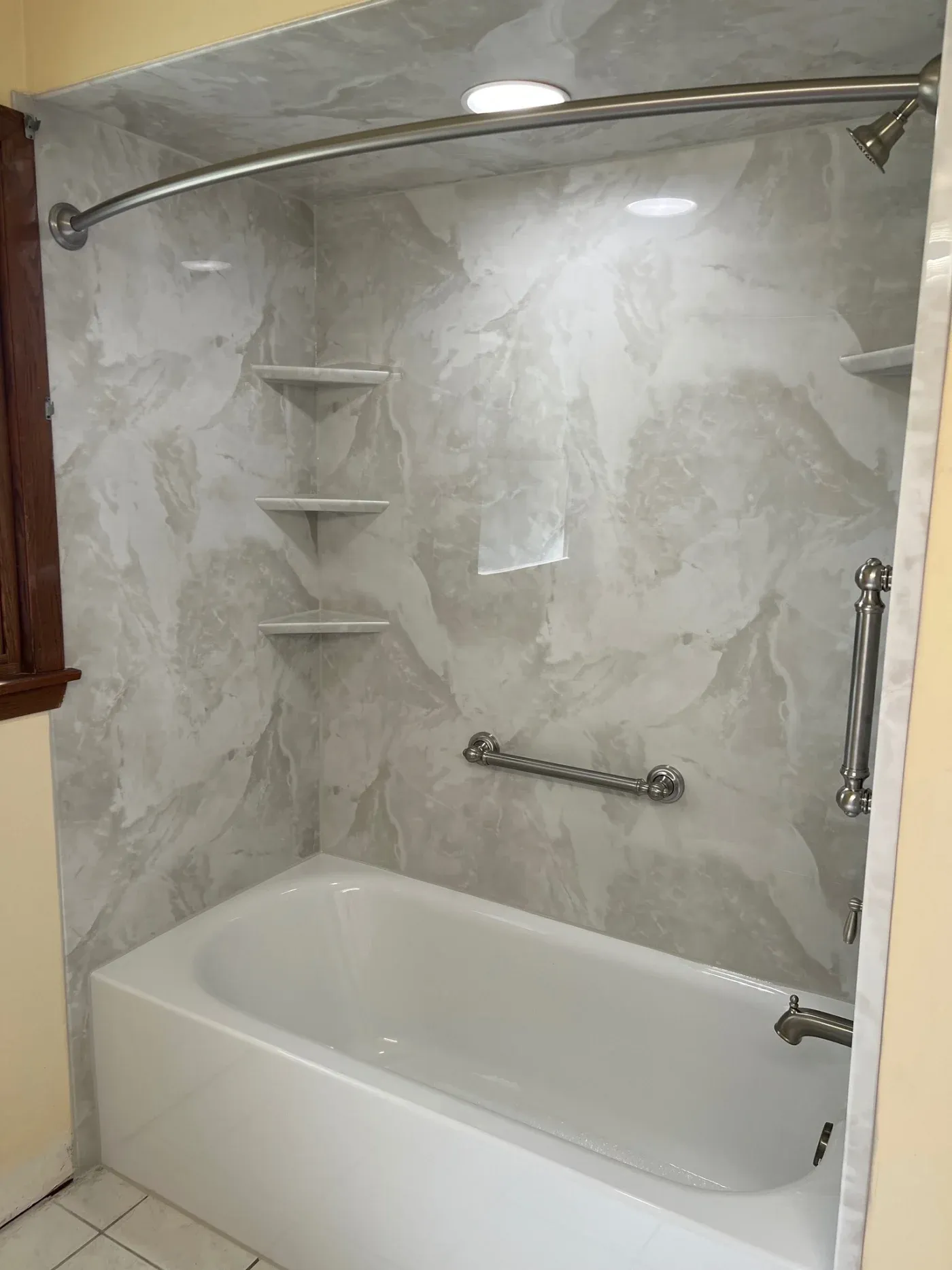 Bathroom shower enclosure with white bathtub, marble-look wall surround, curved chrome rod, showerhead, grab bars, and three corner shelves for storage