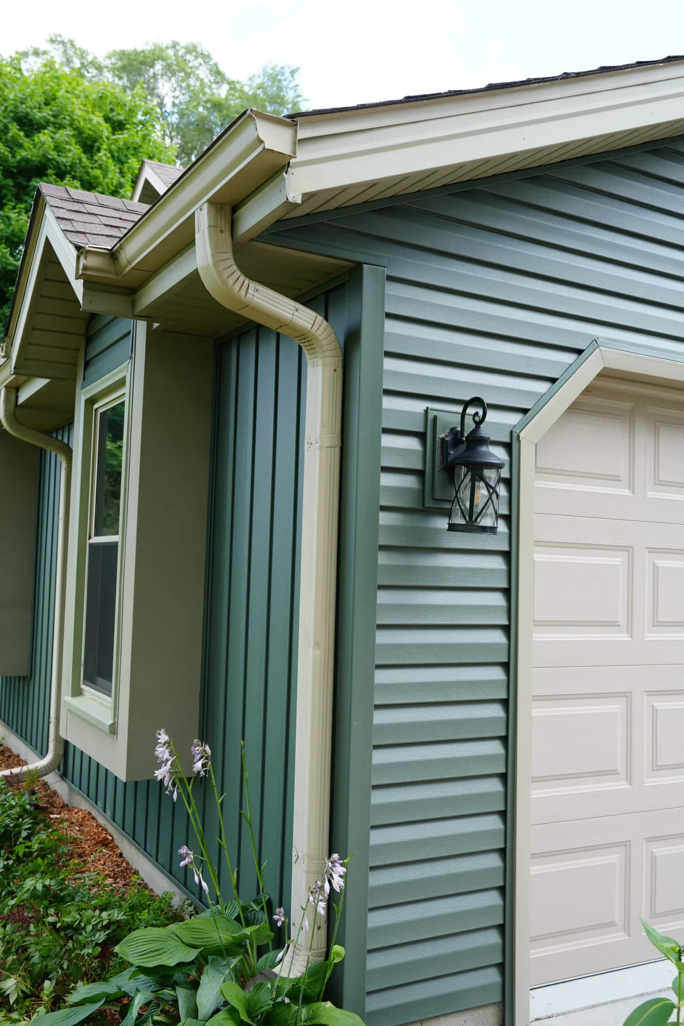 Home exterior showing green siding, white garage door, gutter system, and flowering plants along the foundation