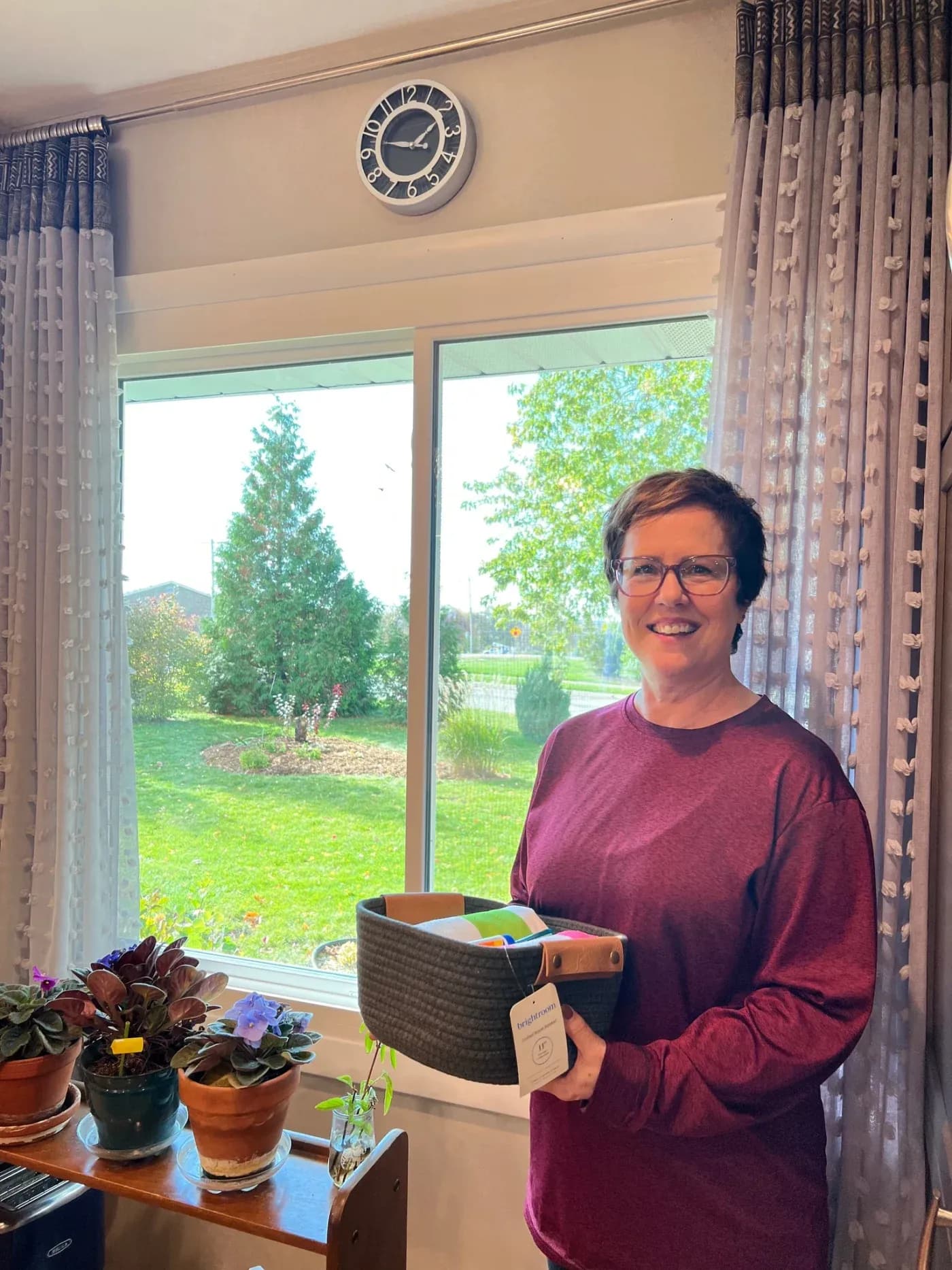 Woman wearing glasses and maroon shirt holding a woven basket with office supplies, standing at a window overlooking a garden with potted plants on the sill