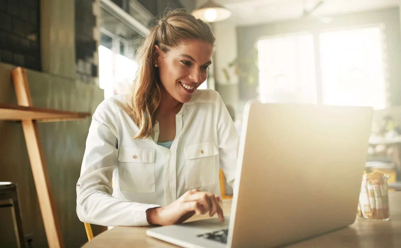 Woman in white shirt smiling while working on laptop at desk in bright office space