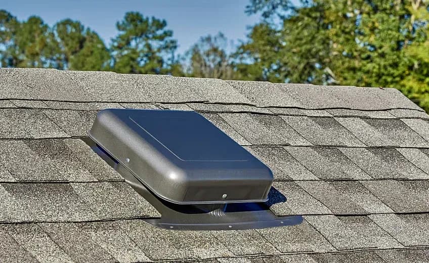 Solar-powered roof ventilation fan installed on asphalt shingle roof with trees in background