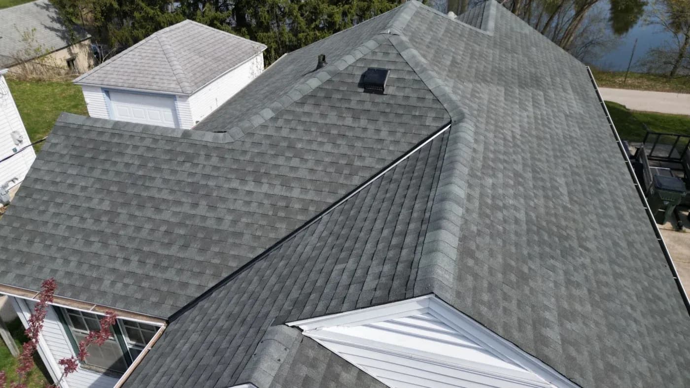 Aerial view of residential house with gray asphalt shingle roof, metal roof panel detail, and white dormers with garage visible in background