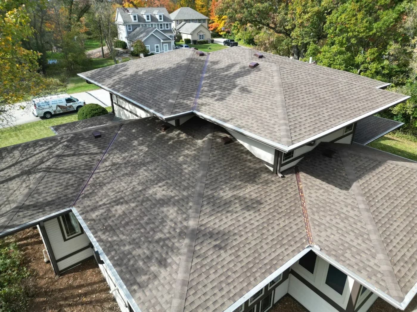 Aerial view of a large house with new architectural shingle roofing installed across multiple roof sections, with measurement lines visible and a contractor's vehicle parked below