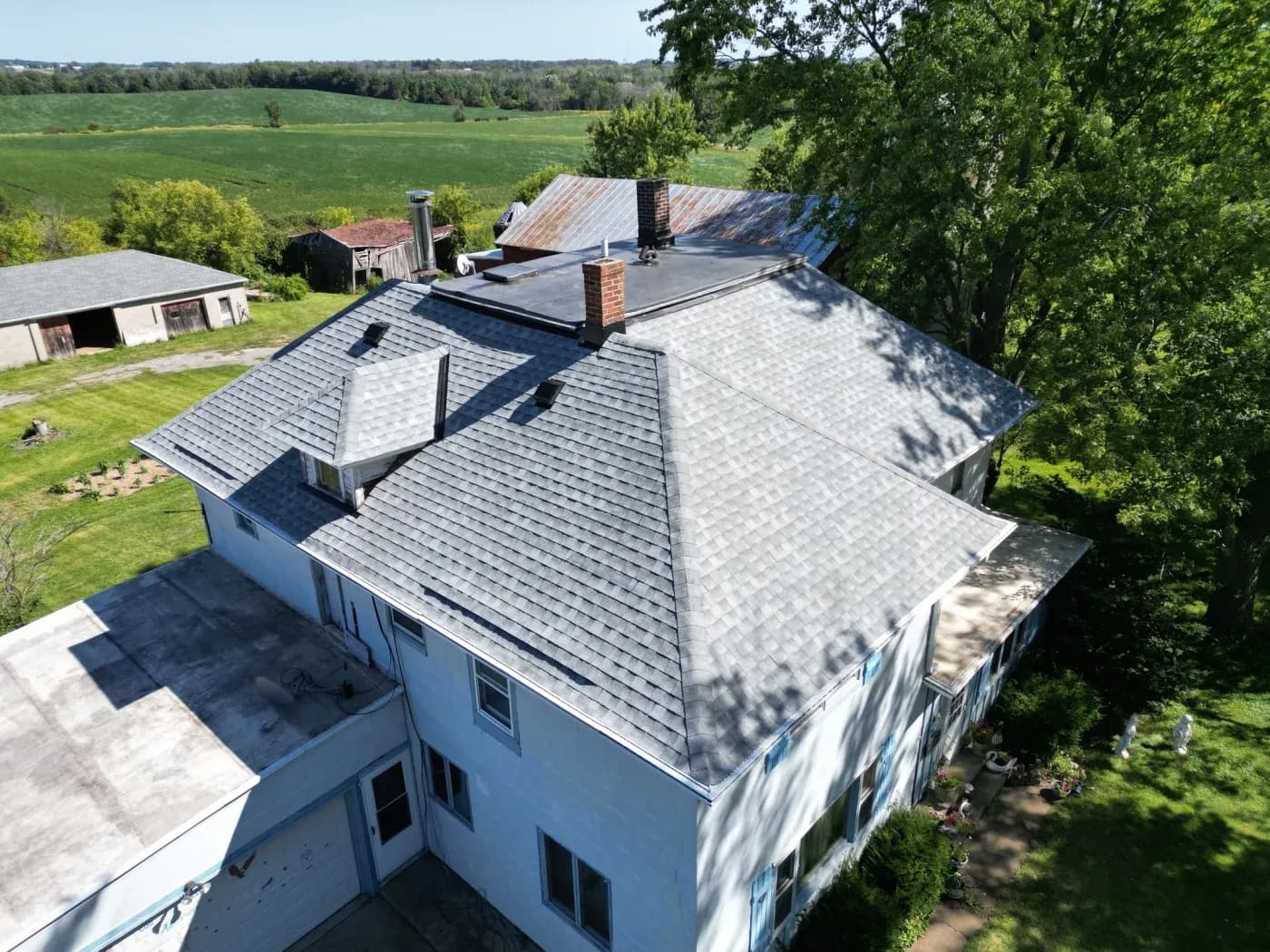 Aerial view of a white farmhouse with new gray asphalt shingle roof, brick chimneys, and attached garage, surrounded by green fields and trees with outbuildings in the background