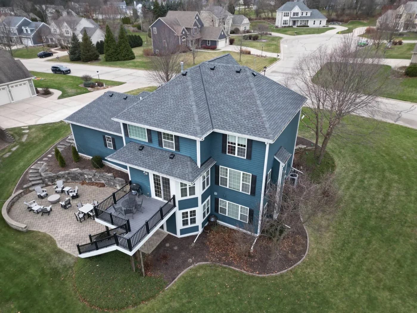 Aerial view of a blue two-story colonial home with gray shingled roof, multi-level deck, patio with seating, and manicured lawn in suburban neighborhood