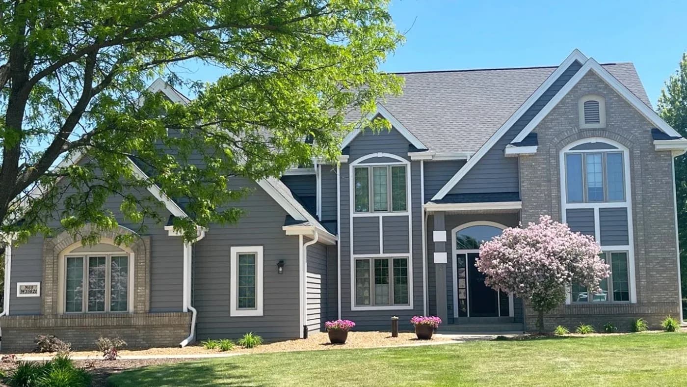 Two-story contemporary home with brick and siding exterior, multiple peaked rooflines, arched windows, flowering magnolia tree, and manicured front lawn with landscaping beds