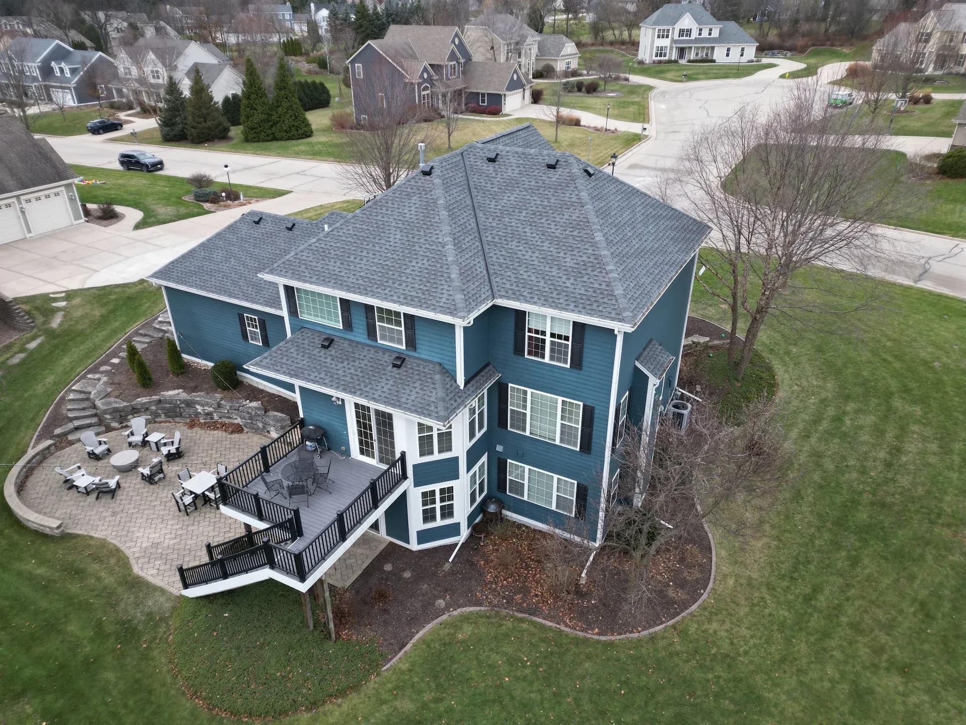Aerial view of a two-story blue house featuring a dark shingle roof, attached deck with black railings, and an extensive paver patio with fire pit seating in a residential neighborhood.