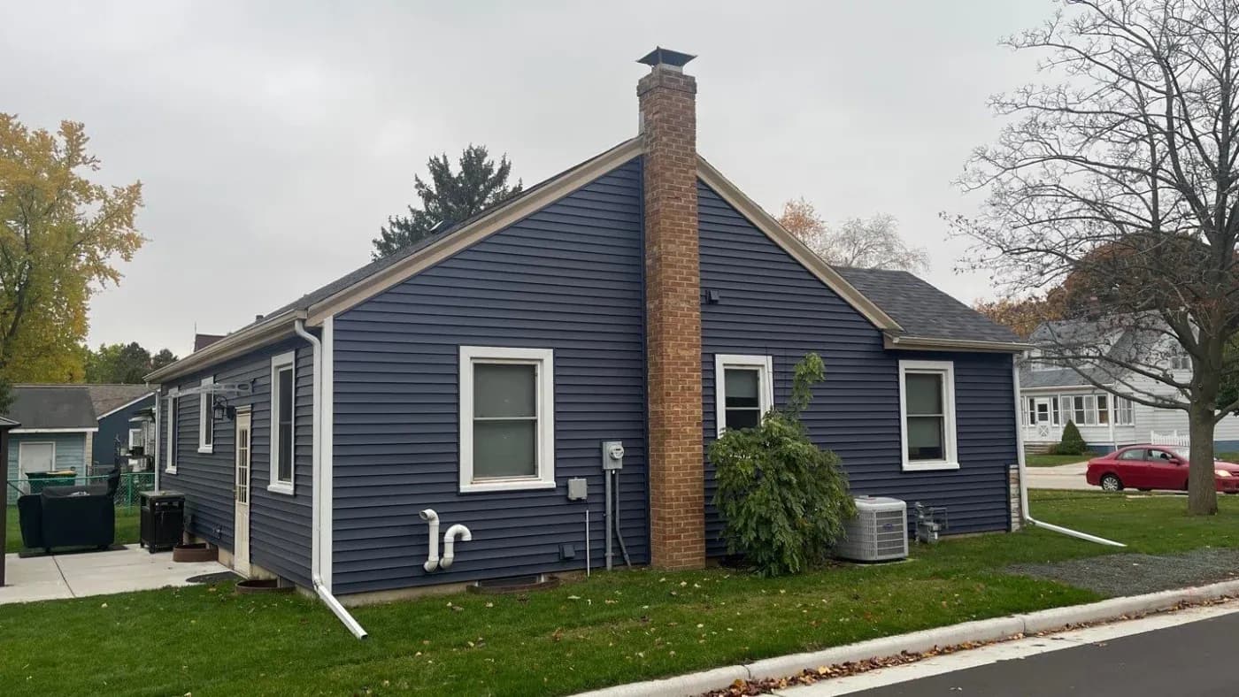 Navy blue single-story house with white trim, brick chimney, three windows, and covered front porch on residential street with fall foliage