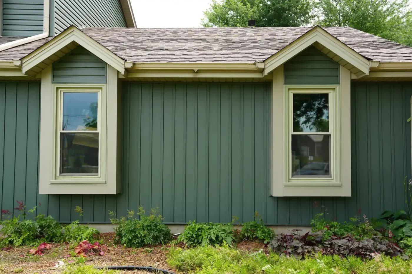 House exterior with green metal siding, two gabled window bays with cream trim, and landscaped garden beds with flowering plants and shrubs below