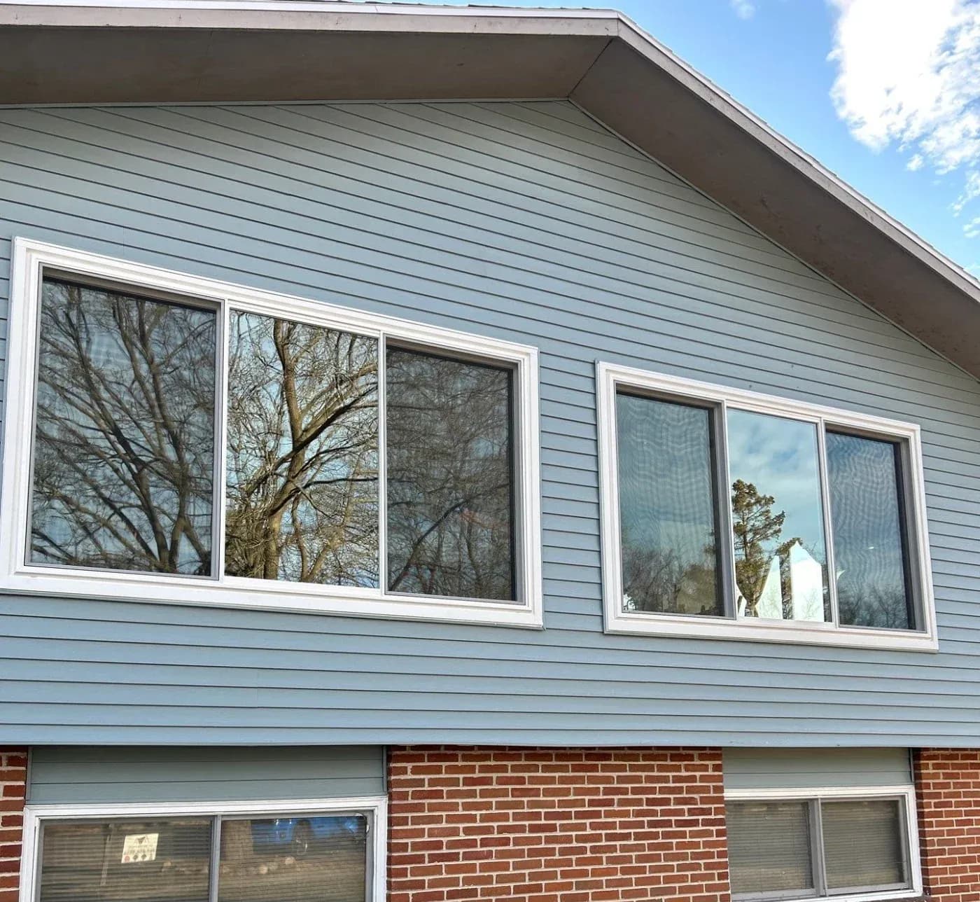 Modern house exterior with light blue horizontal siding and two sets of white-framed windows reflecting bare trees and sky, built above red brick foundation