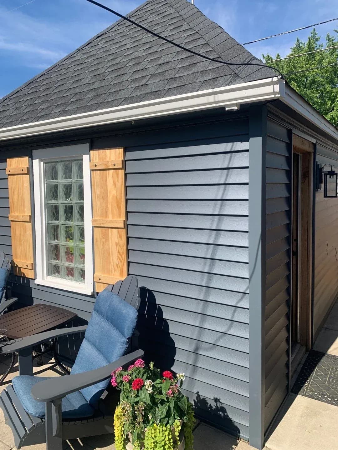 Modern garden shed with white horizontal siding, gray roof, wood shutters, and gridded window, with Adirondack chair and flowering planters in foreground