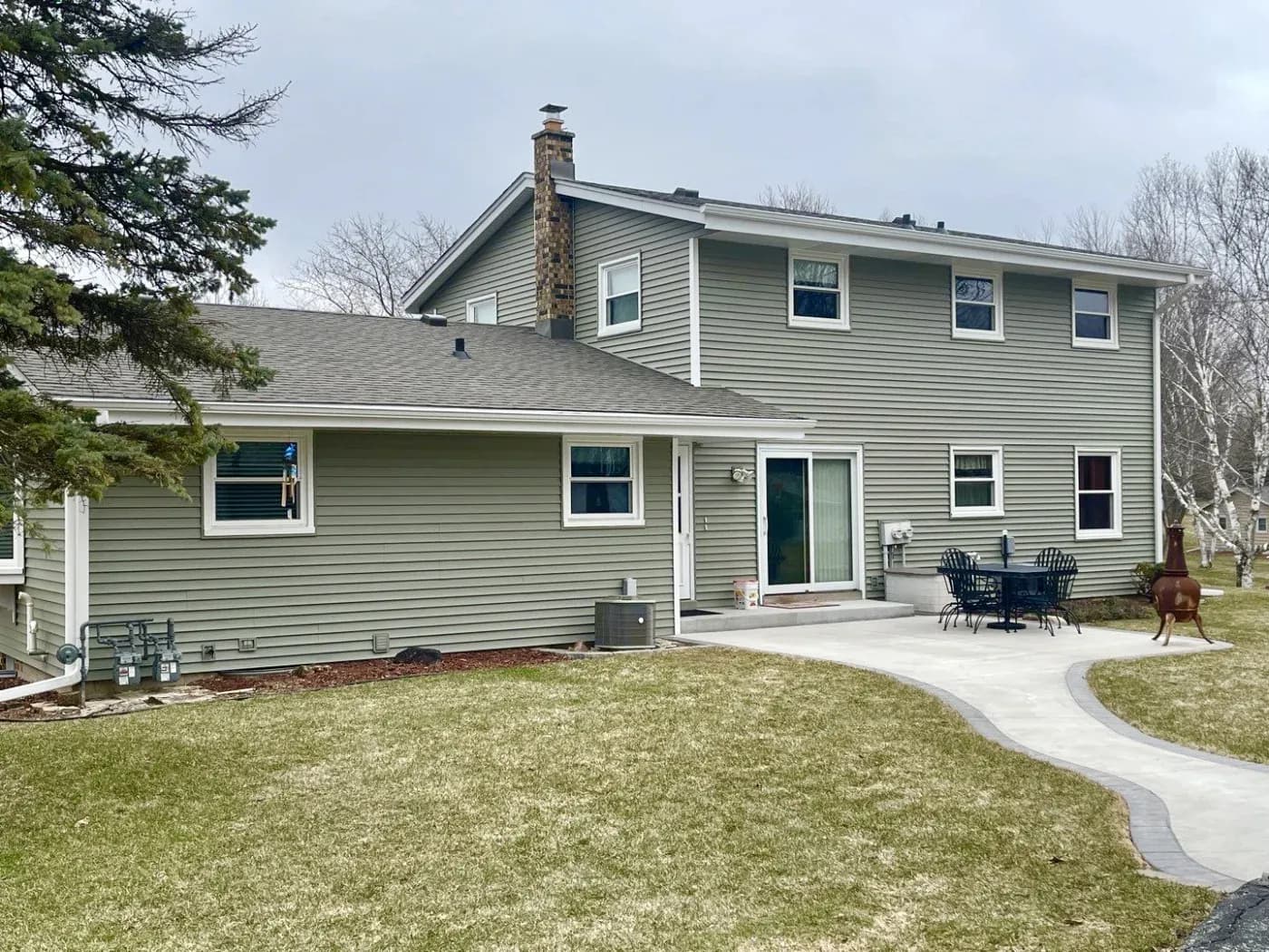 Two-story gray and white residential home with brick chimney, multiple windows, patio with Adirondack chairs, and curved concrete driveway through landscaped yard
