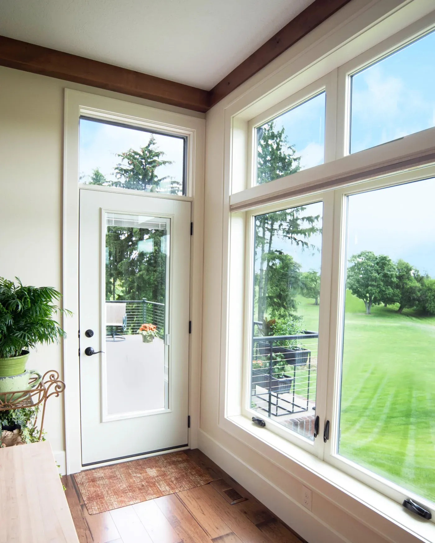 Bright sunroom with white door and large windows overlooking green lawn and trees, wooden beams overhead, potted plants on left side