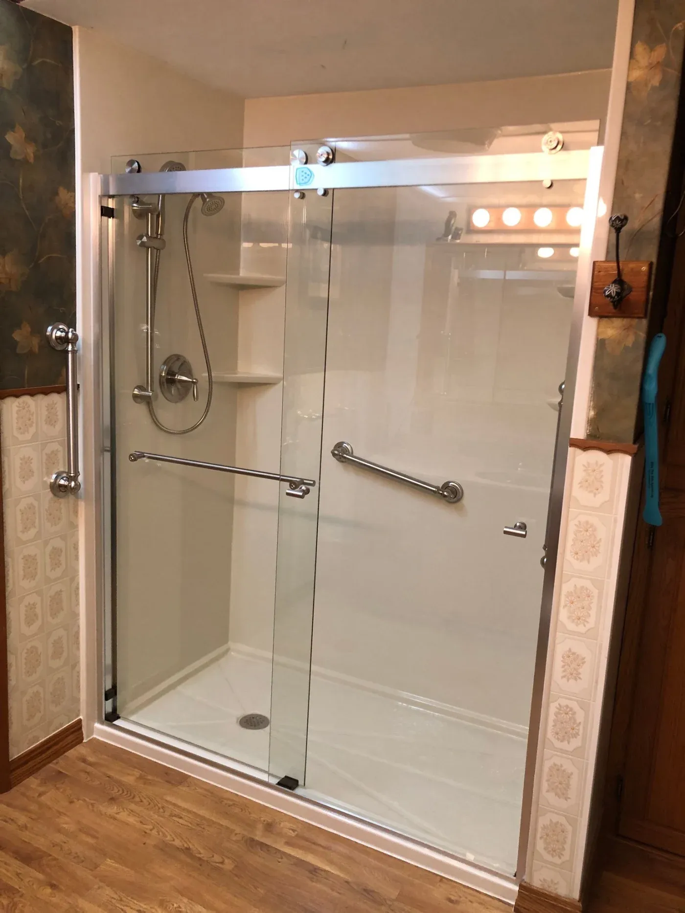 Modern glass shower enclosure with sliding doors, chrome fixtures, handrails, and built-in shelving in a bathroom with decorative tile walls