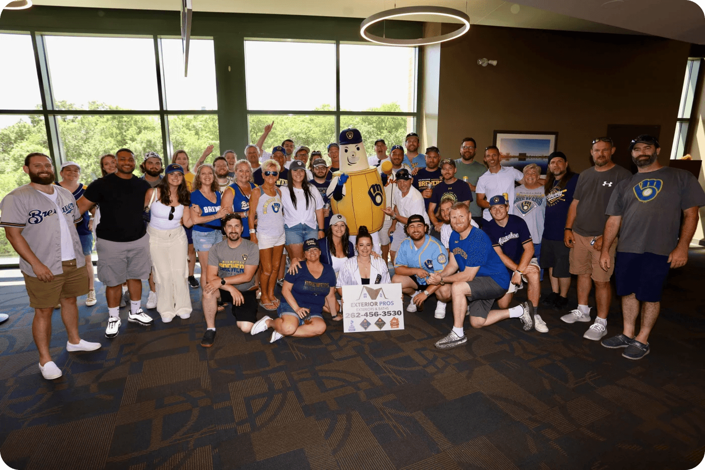 Large group of Milwaukee Brewers fans and staff posing together indoors with the team mascot Bernie, holding a business sign for Exterior Pros