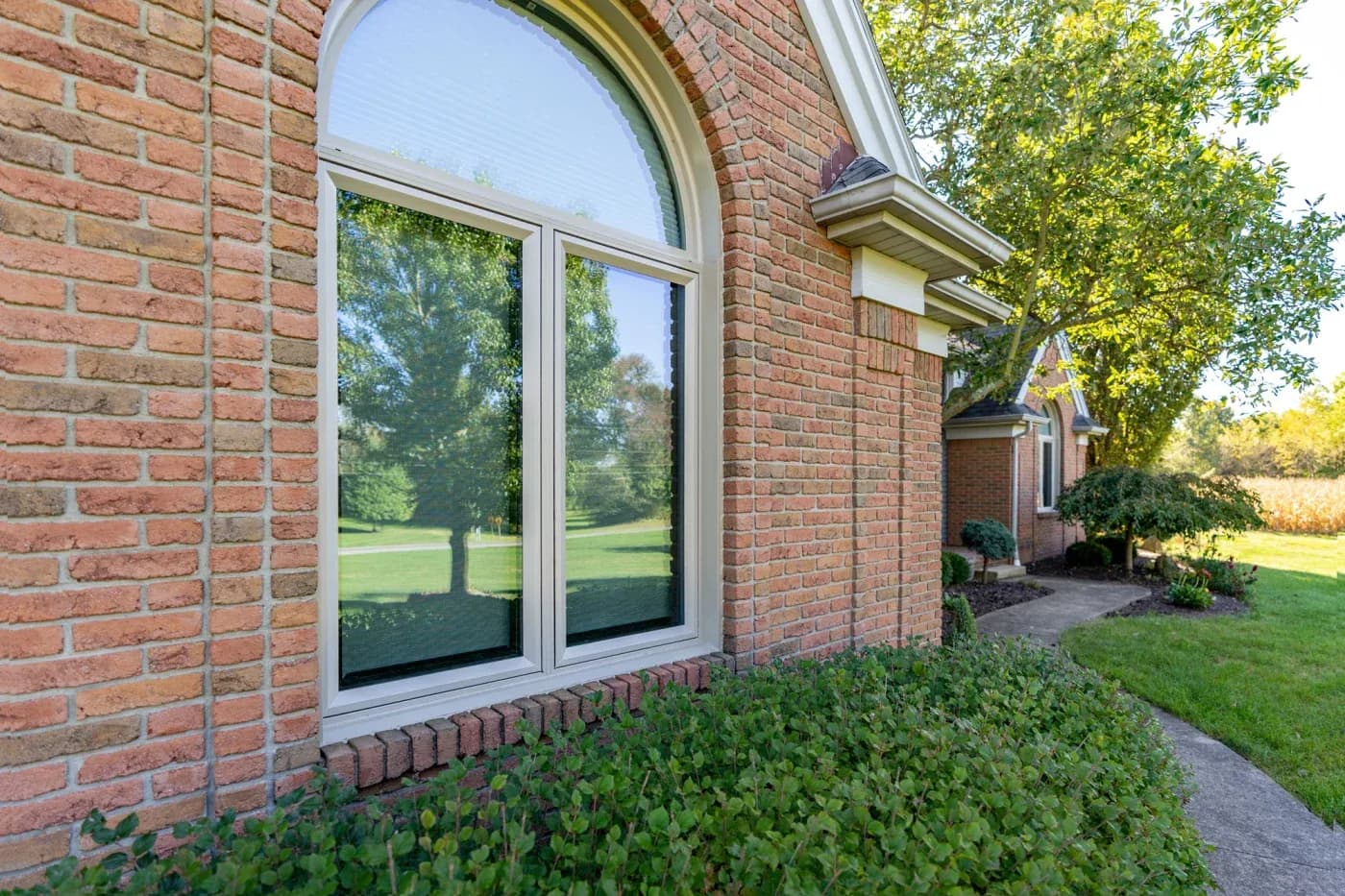 Large arched window with black-framed panes on brick home exterior, reflecting landscaped grounds and trees