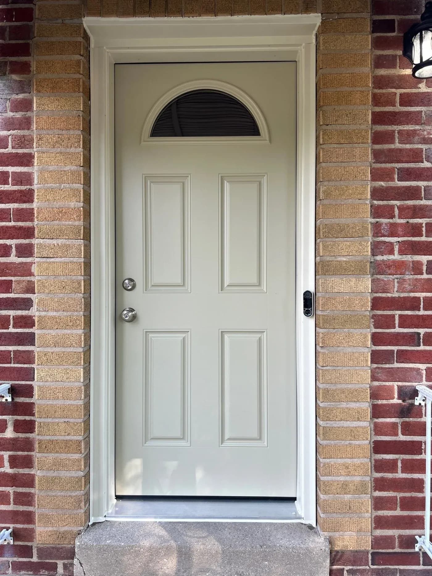 Cream-colored front door with four raised panels and transom window, flanked by red brick columns, with chrome door handles and video doorbell