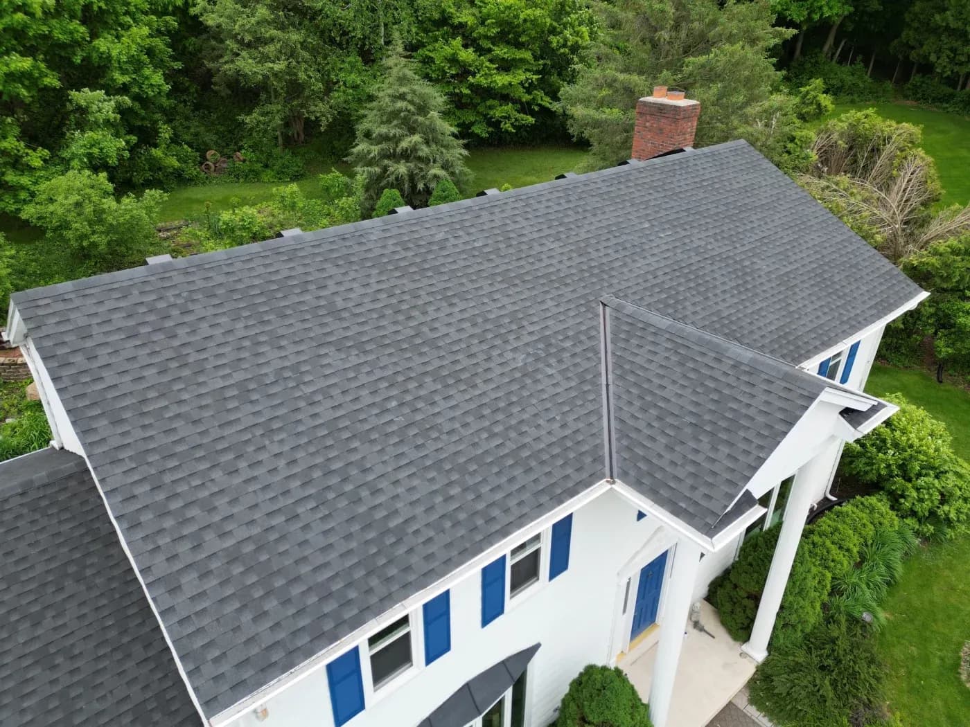 Aerial view of a white house with dark gray asphalt shingles roof and blue shutters, surrounded by mature trees and green lawn