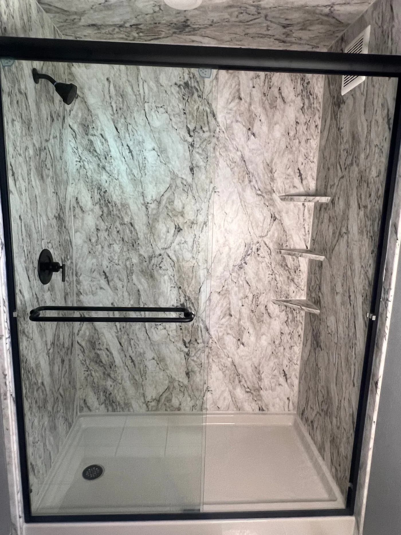 Marble tile walk-in shower enclosure with black framed glass sliding door, shelving, and rainfall showerhead