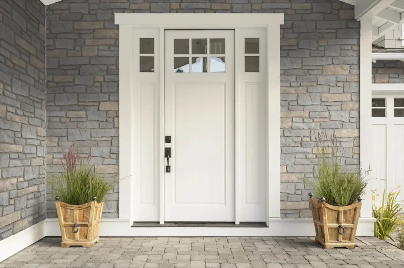 Modern home entrance with white front door featuring glass panels, flanked by sidelights, wooden planters with ornamental grasses on gray stone patio, and gray brick facade