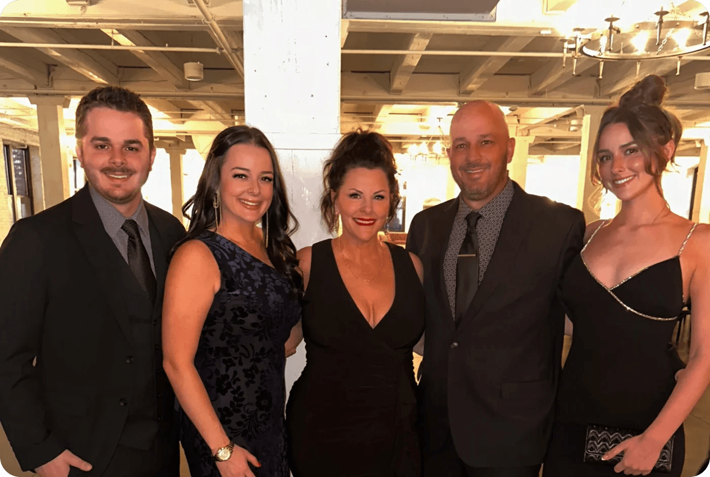 Five people in formal attire posing together at an indoor event with exposed ceiling beams and industrial lighting