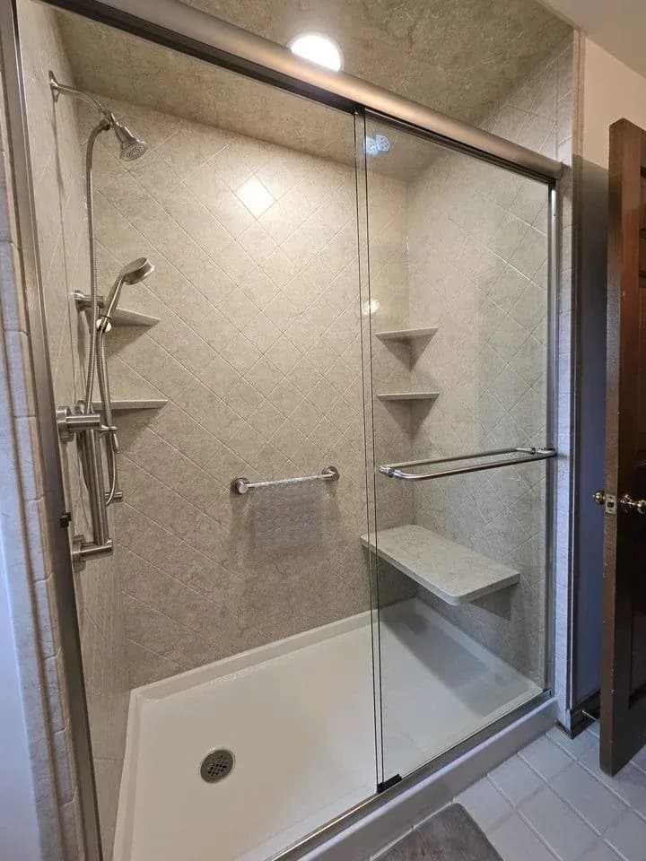 imgi_1_transformative-tub-to-shower-conversions-allenton-wi-exterior-pros-and-baths.webp Interior view of a renovated walk-in shower enclosure featuring beige diagonal diamond-patterned tile surrounds, sliding glass doors with brushed nickel hardware and towel bars, a low-step white shower base, dual showerheads including a detachable hand wand, a built-in shower bench, a horizontal safety grab bar, and corner shelving units.