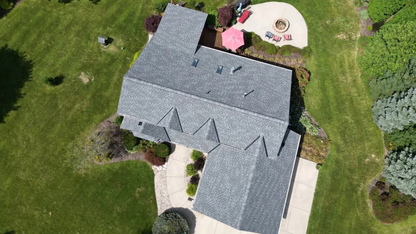 Aerial view of a residential home with gray roof, curved patio with fire pit, pink umbrella, lounge chairs, and manicured lawn surrounded by landscaping