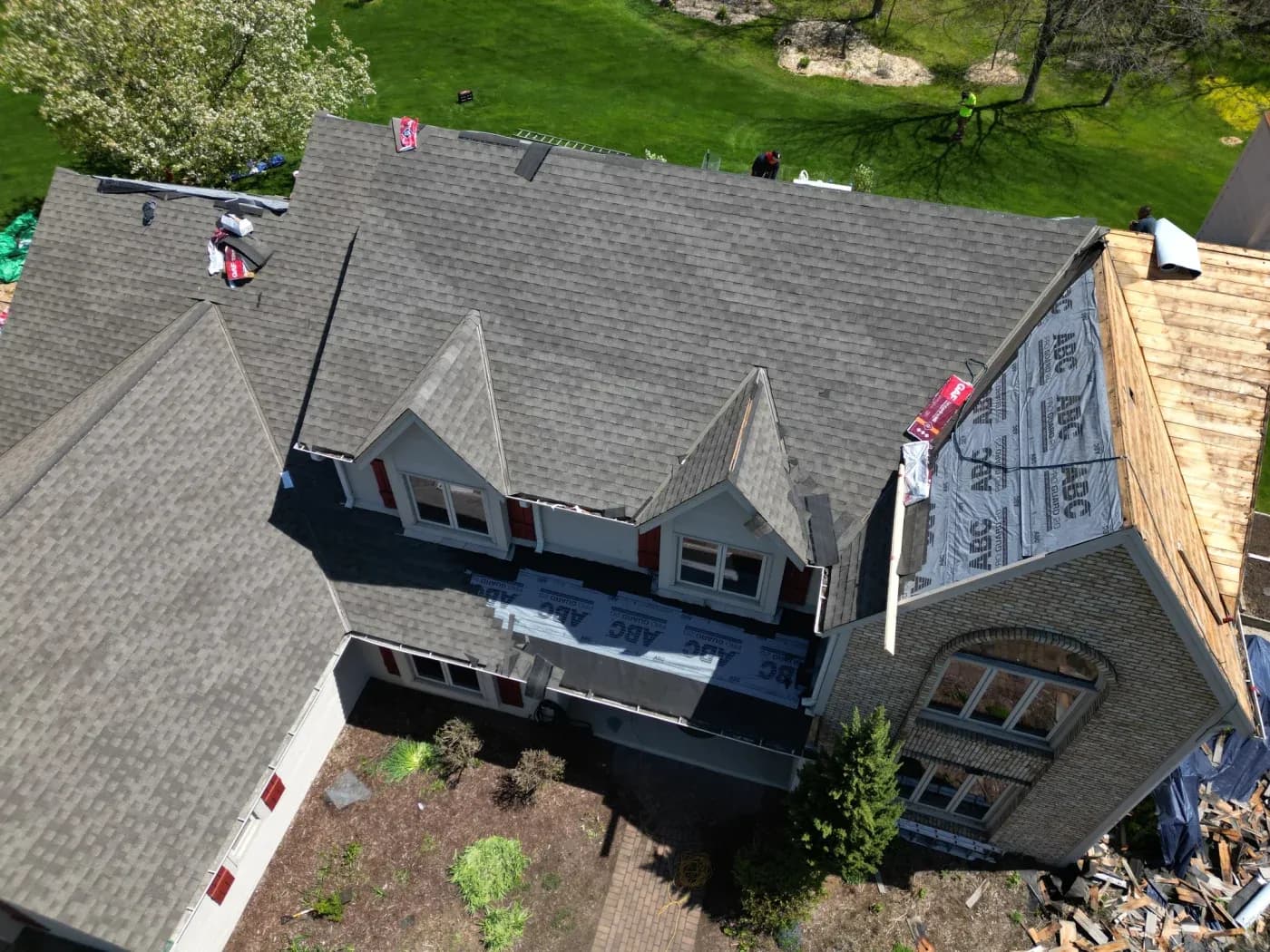 Aerial view of residential roof under construction with gray shingles, dormers, workers, and protective tarping visible from above