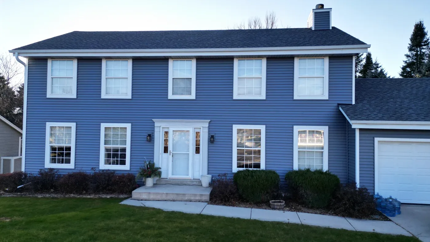 Two-story blue colonial house with white trim, six upper windows, four lower windows, white front door with sidelights, attached garage, concrete walkway, and landscaped shrubs