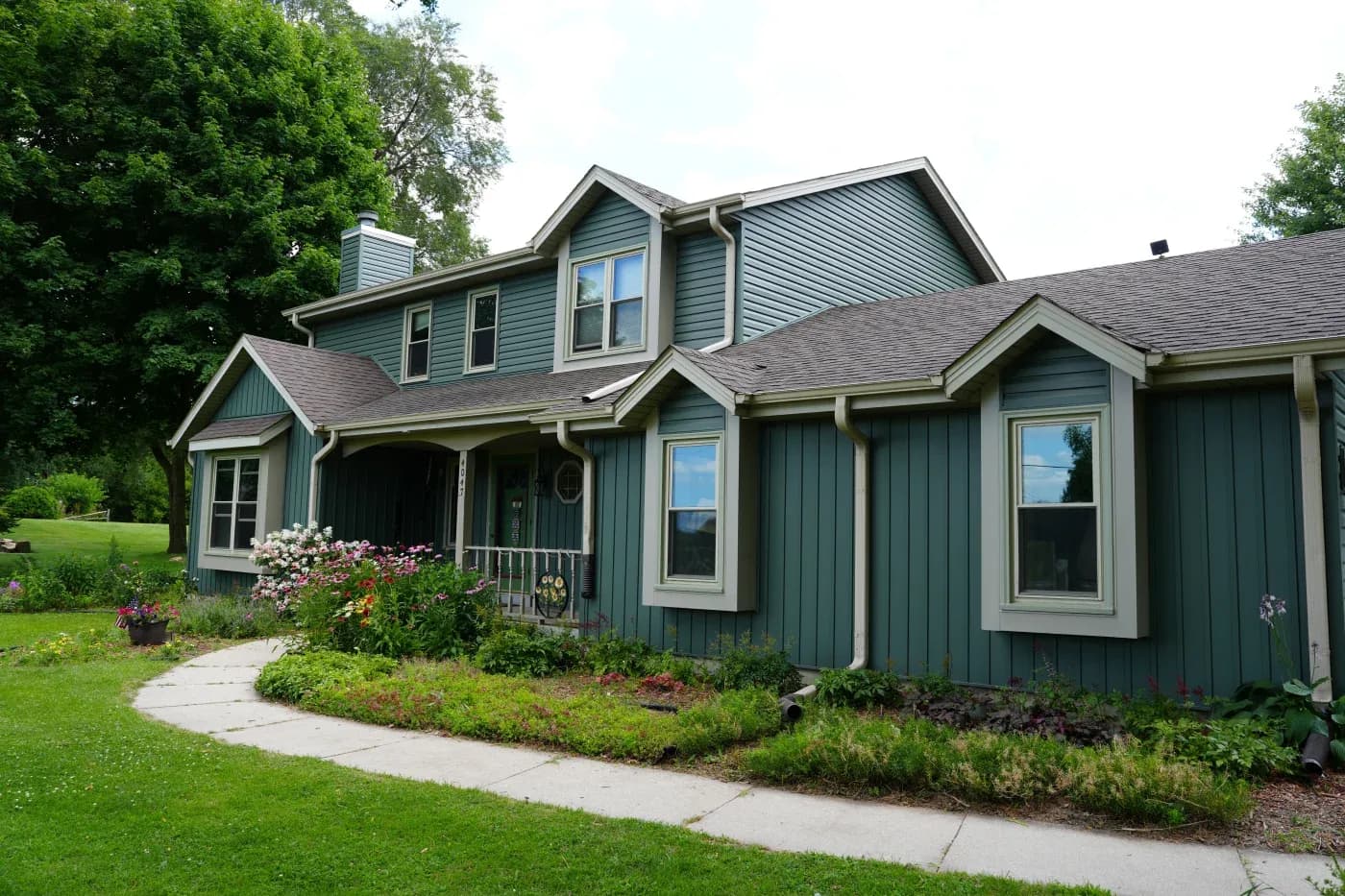 Two-story teal and gray contemporary house with covered porch, flowering garden beds, and curved walkway bordered by mature trees and lawn