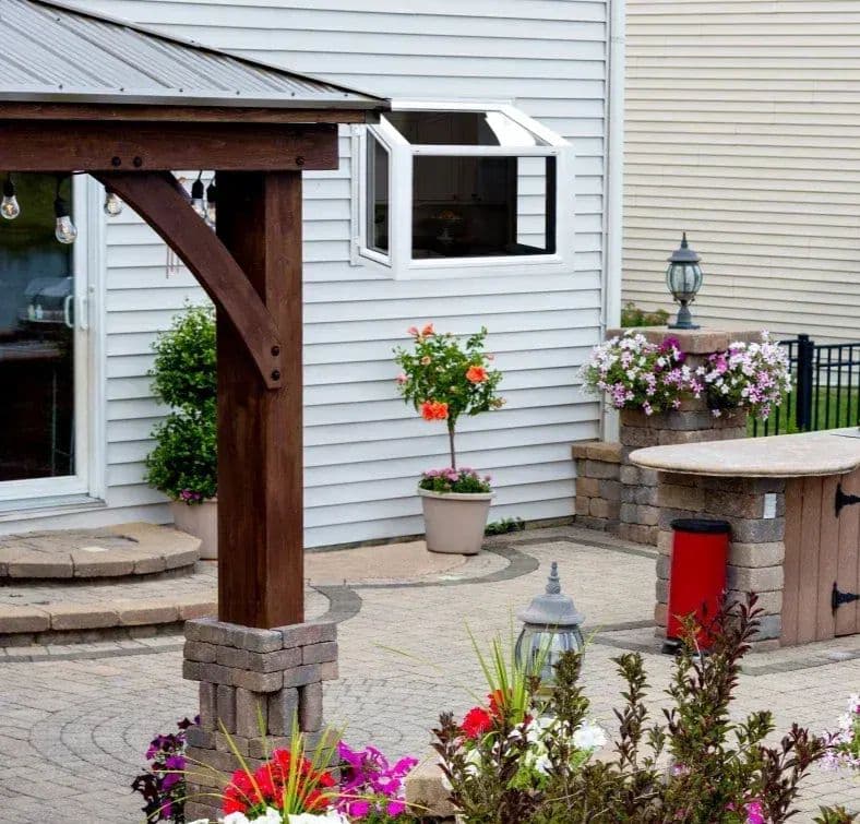 Backyard patio with stone pavers, wooden pergola, potted flowers in red and pink, decorative lamp posts, and seating area attached to white house with dark roof