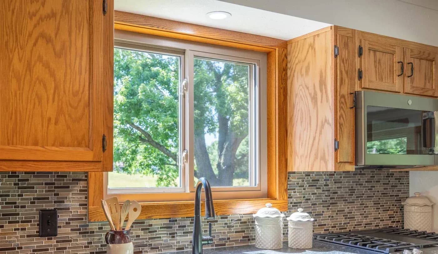 Kitchen with oak cabinetry, brick backsplash, large window overlooking green tree, and black faucet at sink with utensil holder and white canisters