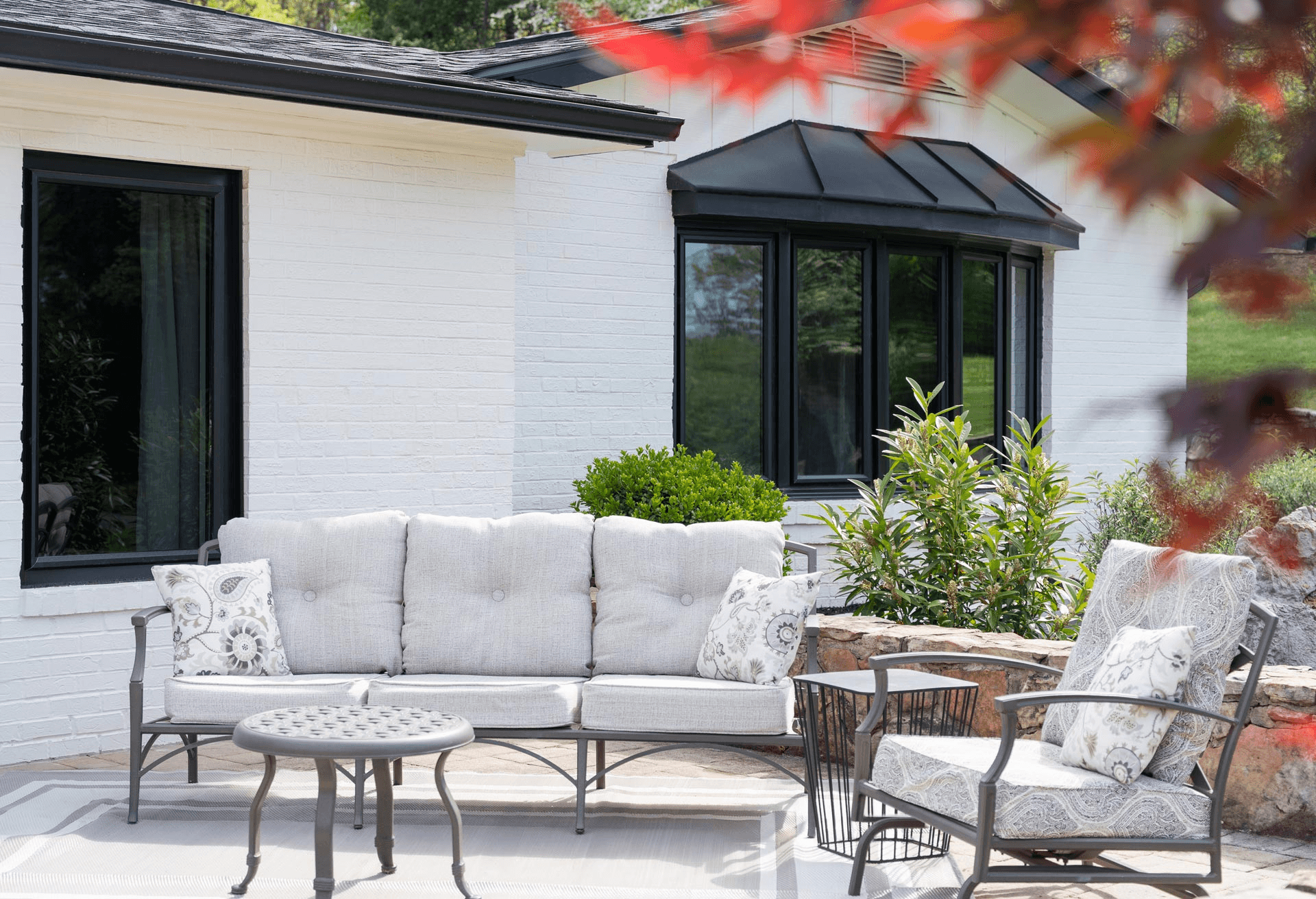 Modern patio with gray upholstered sofa, metal frame chairs with patterned cushions, and round side table in front of white brick house with black-framed windows and skylight, surrounded by landscaping and fall foliage