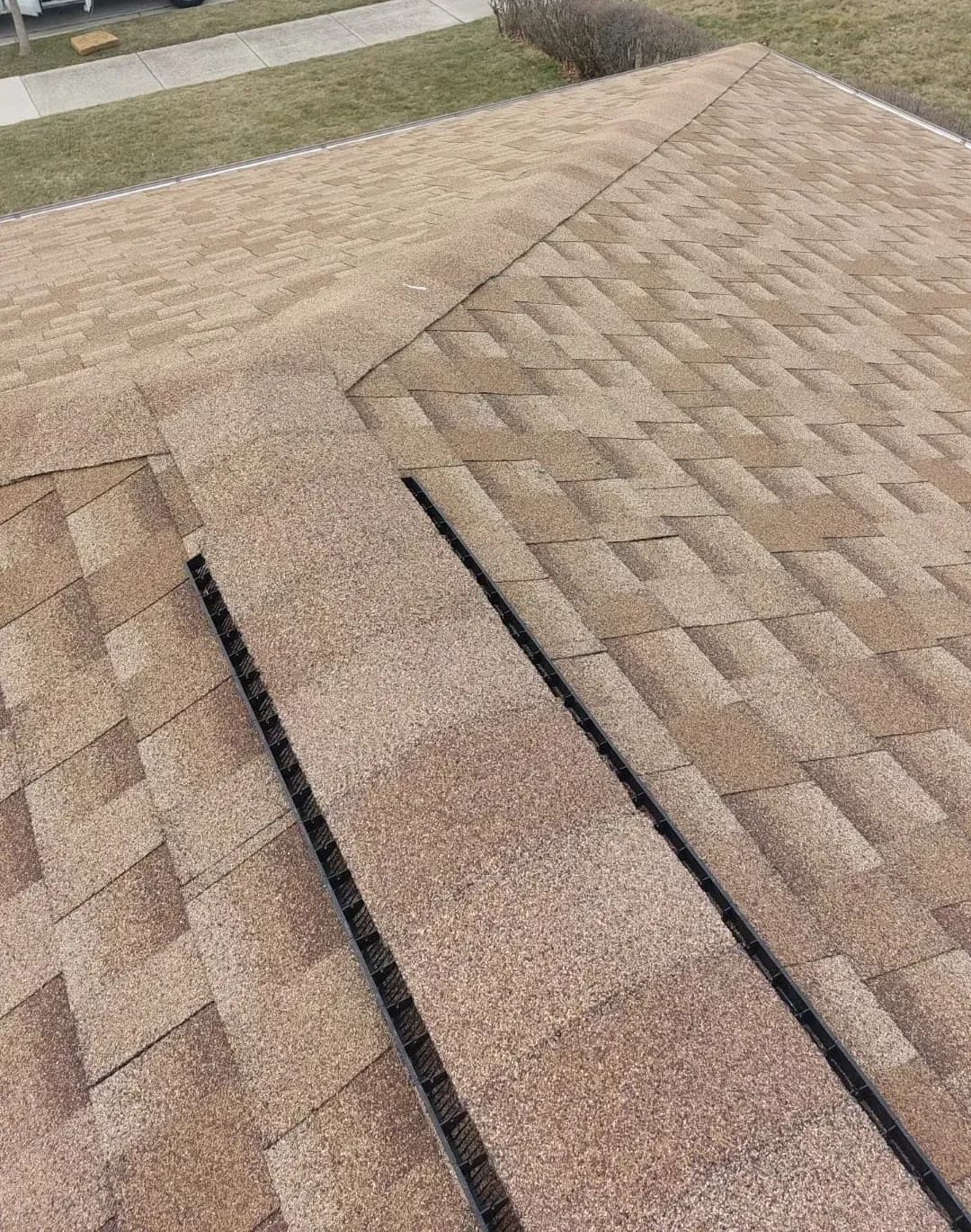 Asphalt shingle roof with two black gutter lines running diagonally across brown architectural shingles in herringbone pattern, residential driveway and lawn visible below