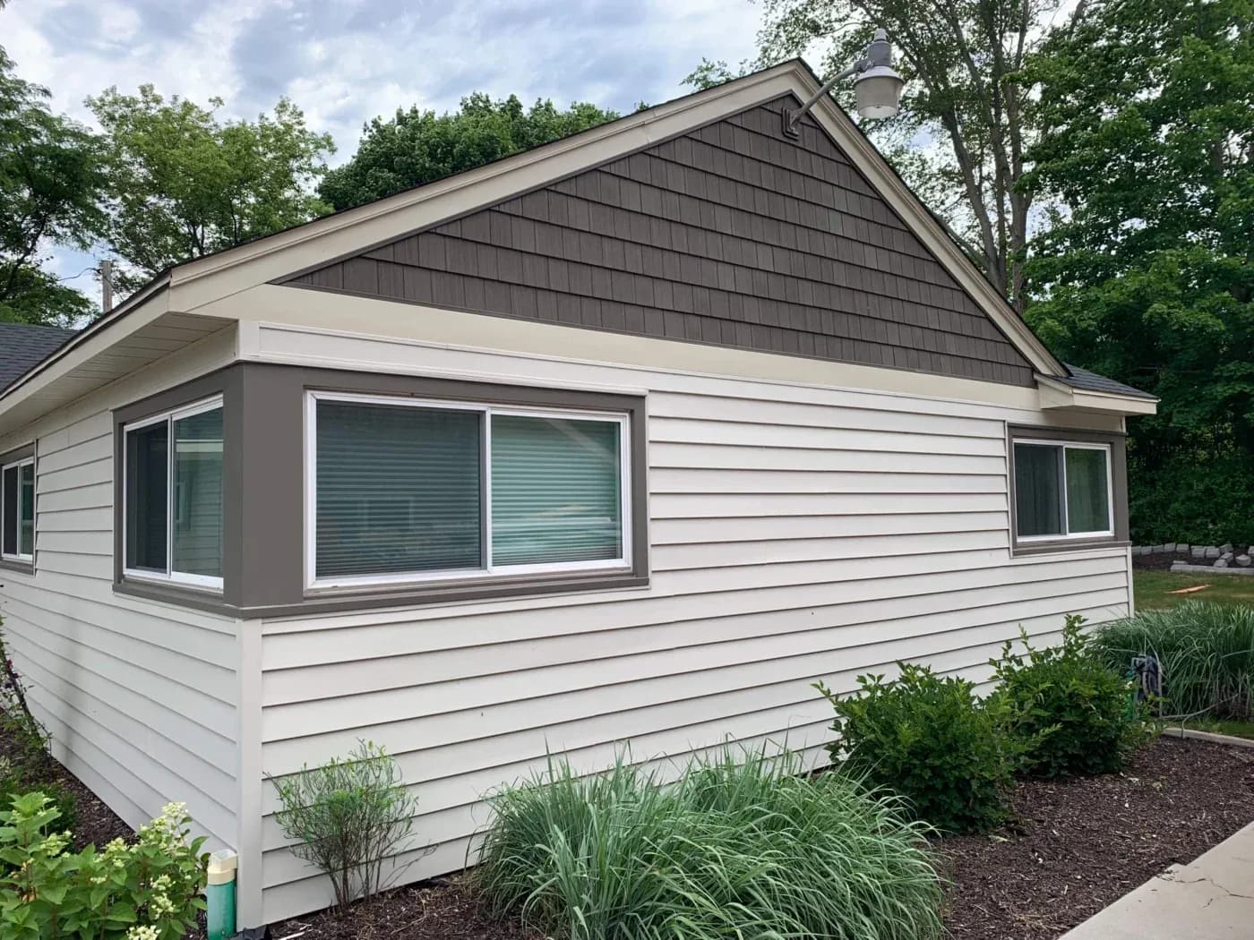 Modern single-story house with white vinyl siding, dark gray gable roof, and large front windows with gray trim surrounded by landscaping and trees