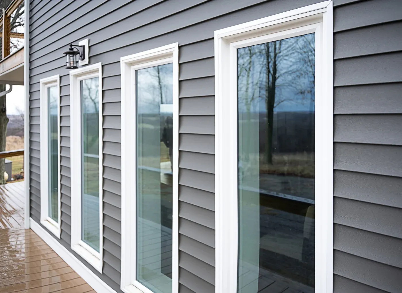 Row of white-framed sliding glass doors on gray house exterior with wooden deck, overlooking bare trees and water view