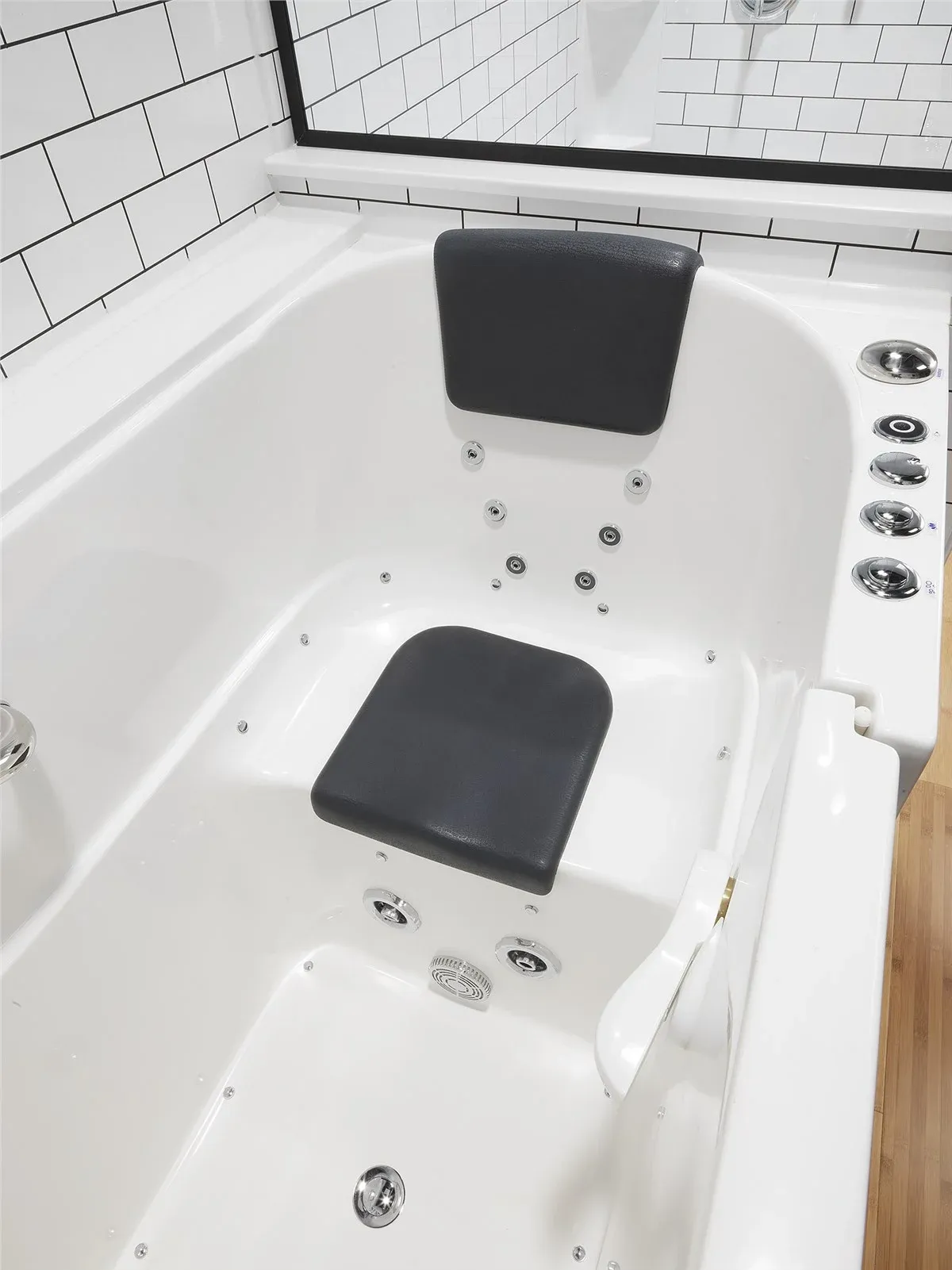Overhead view of a white soaking tub with black cushioned headrest and seat, multiple massage jets, and control buttons on the rim, surrounded by white subway tile walls