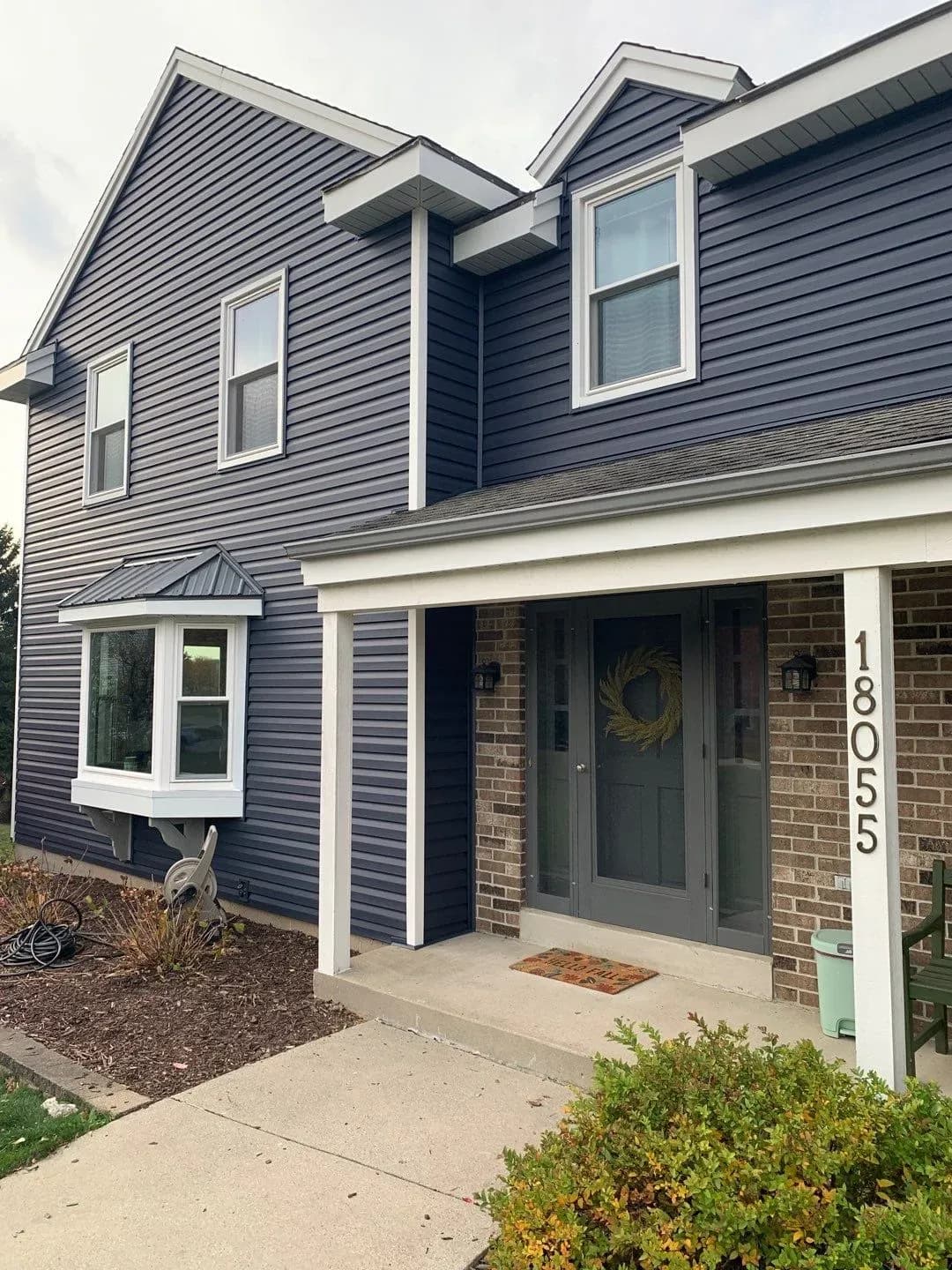 Two-story contemporary home with dark blue horizontal siding, white trim, brick foundation, and gray front door with wreath at address 1805
