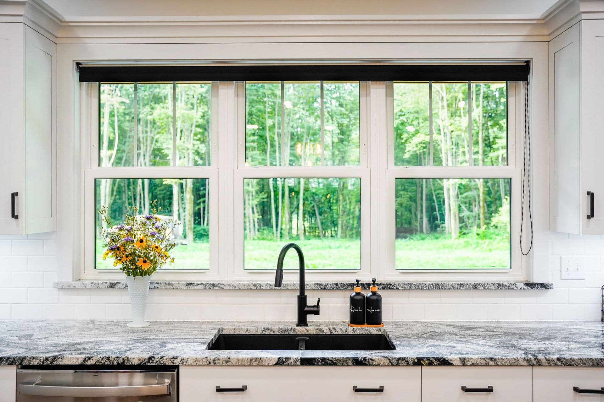 Kitchen sink below three large windows overlooking a forest, with a black faucet, granite countertop, white vase of yellow flowers, and forest view beyond - Provia