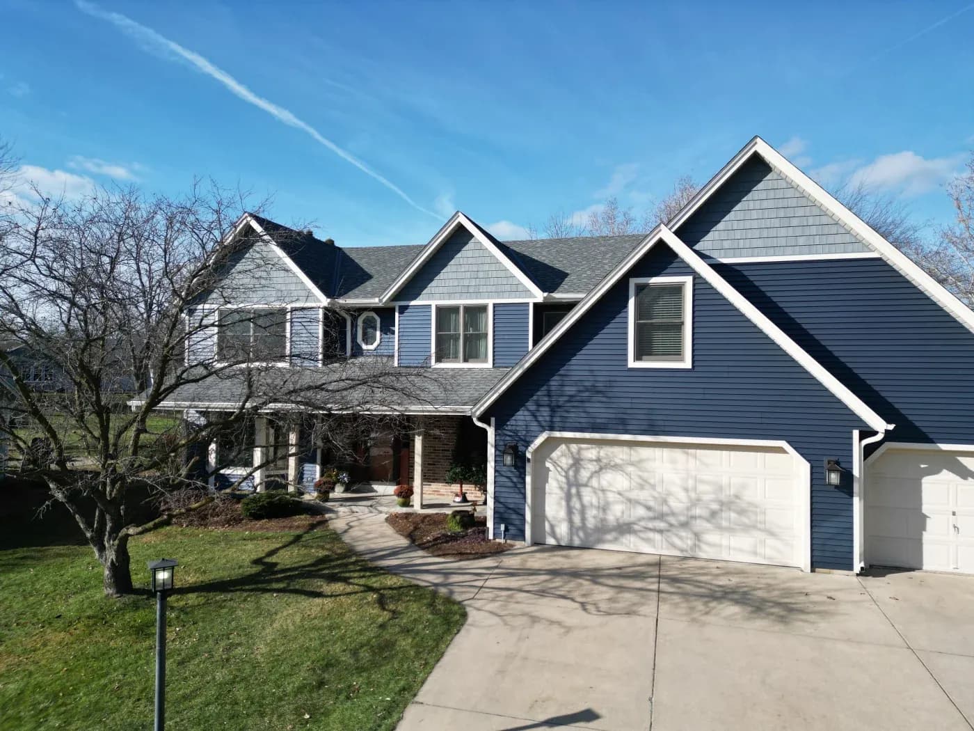 Blue and white two-story suburban home with multiple gabled roofs, white garage doors, front porch, and bare trees in landscaped yard under clear blue sky